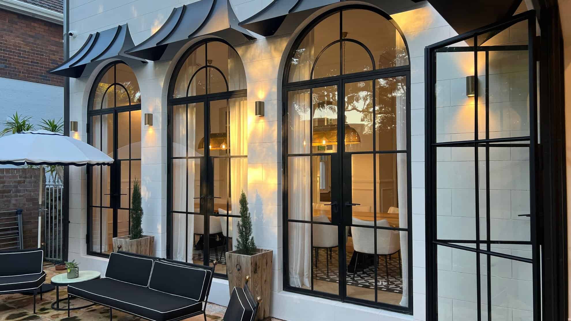 Lit stone facade with arched black-framed doors and outdoor seating at dusk